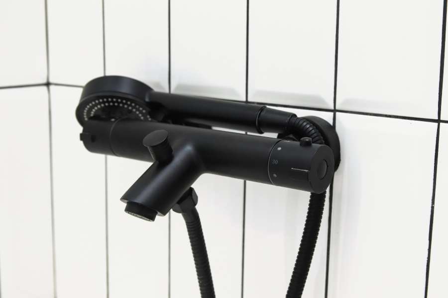 Un mitigeur thermostatique noir mat est fixé sur un mur carrelé blanc avec des joints foncés.